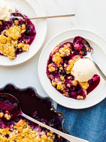 Serving apple and blueberry crumble with scoops of vanilla ice cream on plates.
