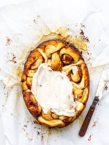 Cinnamon buns on parchment paper being frosted with cream cheese frosting using an offset spatula