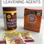 Burgundy and yellow container of Magic baking powder, an orange box of Arm & Hammer baking soda, and packets of Fleischman's traditional yeast
