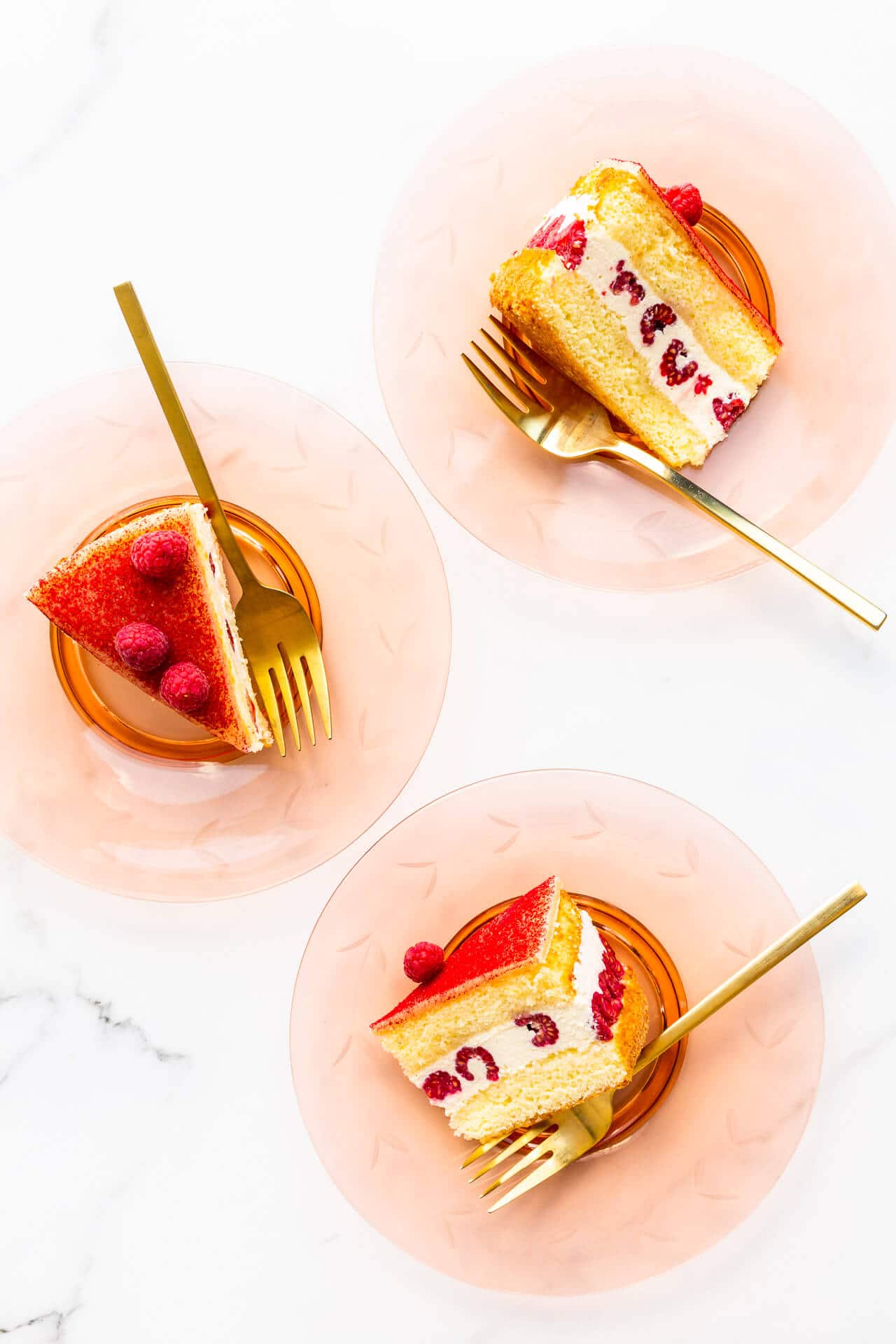 Slices of raspberry and cream layer cake served on glass plates with gold forks.