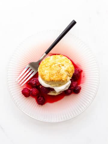 A raspberry shortcake with homemade biscuit, whipped cream, and berries on a pink plate with a fork with black matte handle