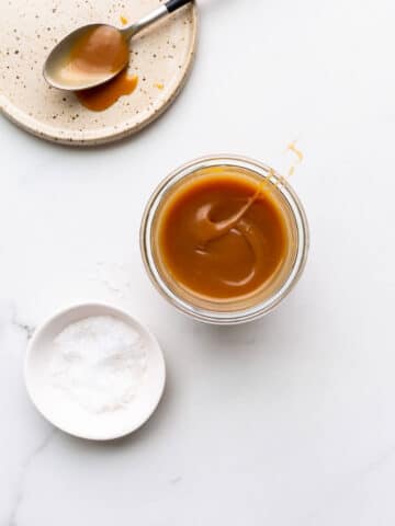 Homemade salted caramel sauce in a jar with a little pinching bowl of sea salt and a ceramic plate with a spoon with salted caramel on it