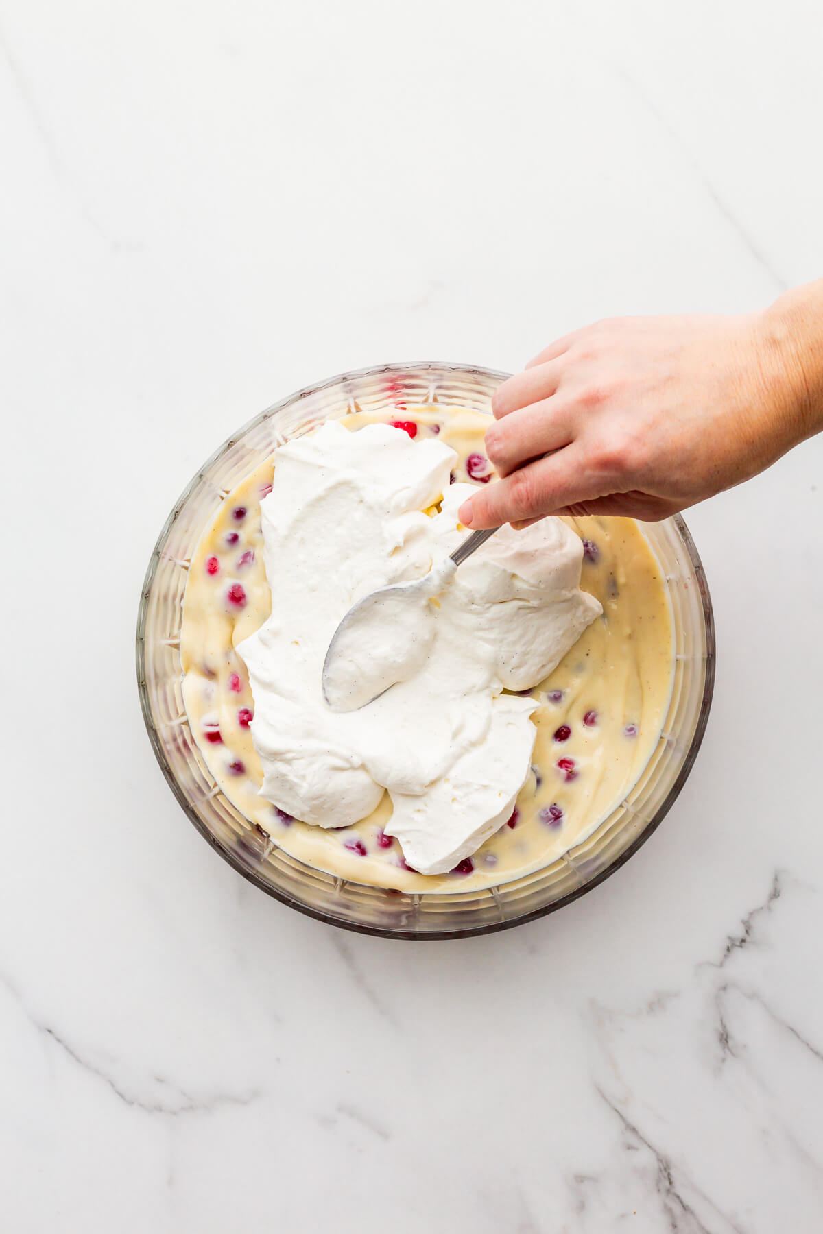 Spreading whipped cream over a layer of pastry cream to make trifle in a glass bowl.